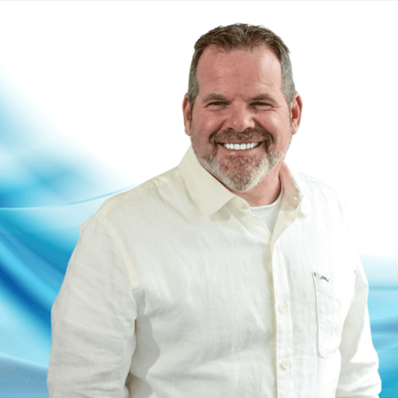 Portrait of a smiling, middle-aged man with a gray beard wearing a cream button-down shirt against a white and blue abstract background.