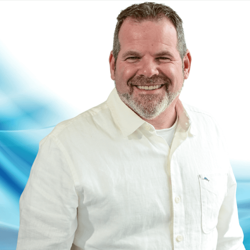 Portrait of a smiling, middle-aged man with a gray beard wearing a cream button-down shirt against a white and blue abstract background.